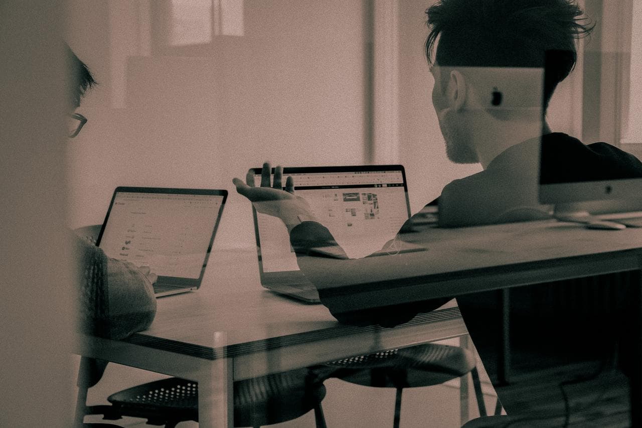 Two people sit at a table working on laptops, discussing something on their screens. The scene is viewed through a glass partition, giving the image a muted, reflective look